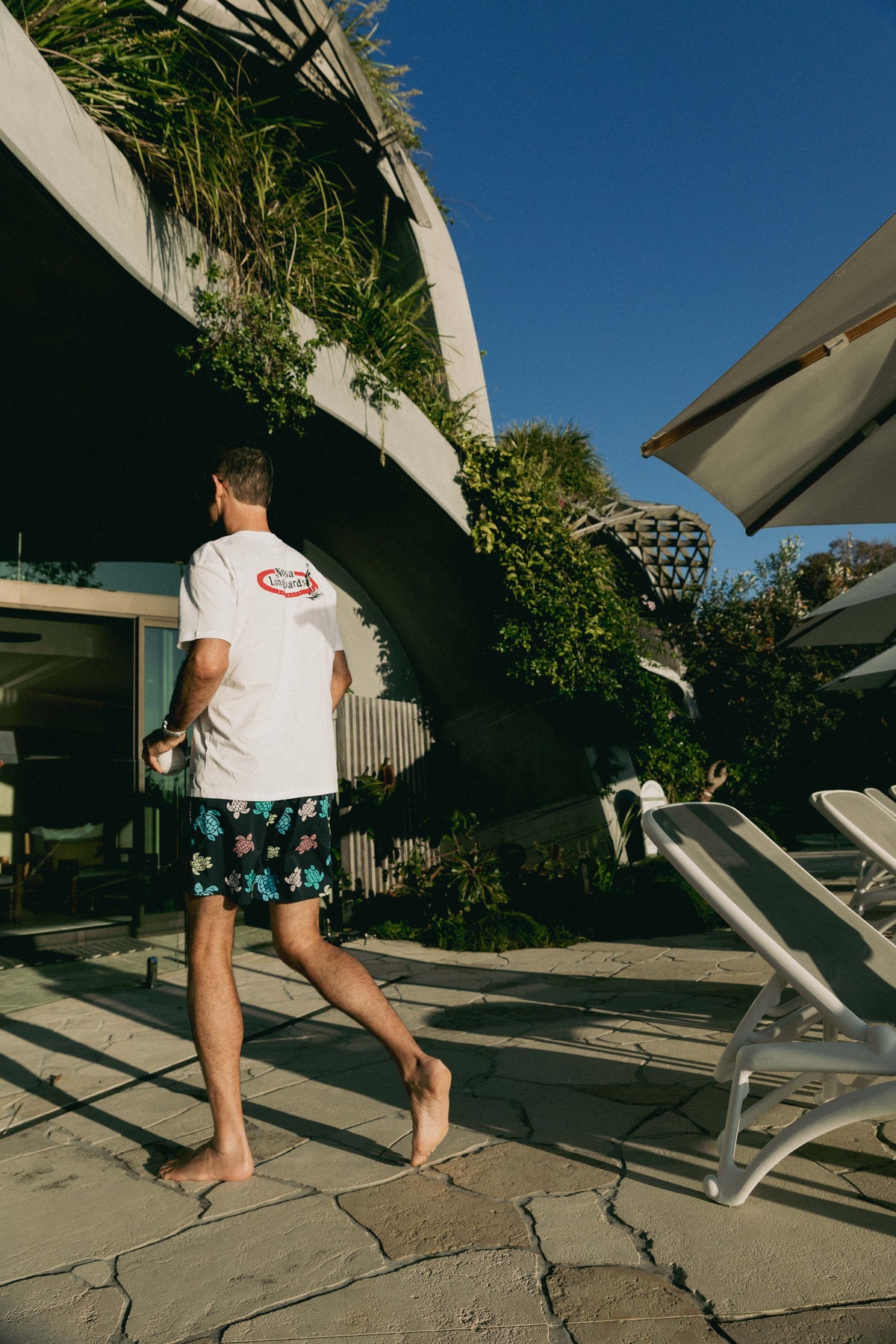 Rear view of NL Original Tee – White worn walking beside the pool at Domic, Sunshine Beach
