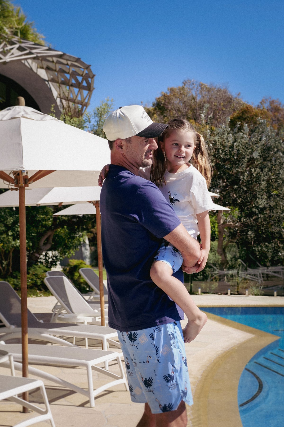 NL Kids Scrub Club Tee – White worn while being held by an adult poolside at Domic